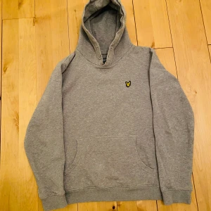 Grå hoodie från Lyle & Scott - Tjena, jag säljer denna gråa hoodien från Lyle & Scott med deras ikoniska logga på bröstet. Tröjan har en klassisk känguruficka och ribbade muddar vid ärmslut och nederkant. Perfekt för en avslappnad stil. Säljer den för 250kr nypris 500-600kr