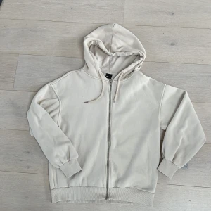 Beige hoodie från Gina Tricot - Säljer en stilren beige hoodie från Gina Tricot i storlek XS. Den har dragkedja framtill och en praktisk huva med dragsko. Perfekt för en avslappnad look.