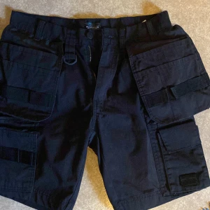 Arbetsshorts storlek S - Säljer ett par svarta arbetsshorts från Bluewear med flera praktiska fickor och bälteshällor. Perfekta för en aktiv dag utomhus. Shortsens robusta design gör dem idealiska för arbete eller fritid. De är i storlek S eller 46 och ganska smala i passformen. 