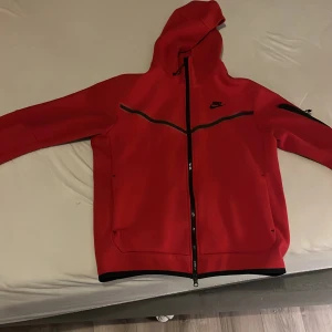 Röd tech fleece hoodie från Nike - Snygg röd hoodie från Nike med dragkedja och huva. Jackan har svarta detaljer och en ficka på ärmen. Lite sönder vid ena fickan men inget som syns jätte mycket. Perfekt för en sportig look. Storlek m men passar s. Nypris 1250kr