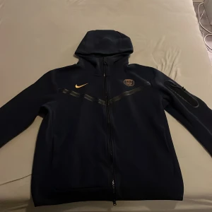Mörkblå psg tech fleece hoodie från Nike - Snygg mörkblå hoodie psg tech fleece från Nike. Köpt på jd sport, som ny bara använd 4 gånger nypris ca 1500. Storlek L men passar M