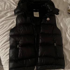 Moncler bromens väst. Moncler Väst - Snygg svart dunväst från Moncler med dragkedja och hög krage. Perfekt för kyliga dagar när du vill hålla värmen utan att kompromissa med stilen. Västen har praktiska fickor med dragkedjor och Monclers ikoniska logga på bröstet. Sjukt fint skick. Rök/djurfri
