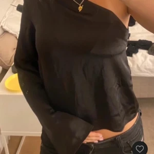 Svart oneshoulder topp - Snygg svart oneshoulder topp med en elegant och stilren design. Perfekt för en kväll ute eller en speciell tillställning. Toppen har en lös passform och är tillverkad i ett glansigt material som ger en lyxig känsla.