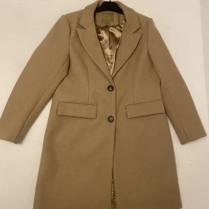 Beige kappa från Amisu - Elegant beige kappa från Amisu med klassisk design och knappar framtill. Perfekt för höst och vår, med långa ärmar och stilrena fickor. En tidlös favorit i garderoben.