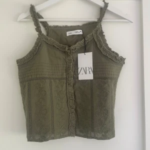 Olivgrön topp från Zara - Snygg olivgrön topp från Zara med dekorativa spetsdetaljer och volangkant. Toppen har smala axelband och ett broderat mönster framtill. Perfekt för varma dagar.