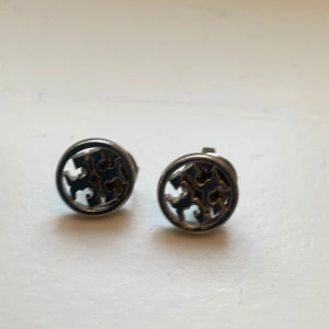 Miller Stud örhängen från Tory Burch - Snygga och stilrena Miller Stud örhängen från Tory Burch i silverfärg. Dessa örhängen har en rund design med det ikoniska Tory Burch-mönstret. Perfekta för att ge en elegant touch till din outfit.