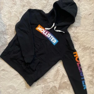 Svart hoodie från Hollister - Säljer en svart hoodie från Hollister med färgglatt logotryck på bröstet och ärmen. Den har en justerbar huva med vita snören och en praktisk magficka. Perfekt för en avslappnad stil.