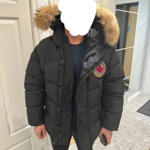 Svart dunjacka från Dsquared2 - Säljer en svart dunjacka från Dsquared2 med en stor, fluffig pälsfodrad huva. Jackan har en broderad logotyp på bröstet och stängs med dragkedja. Perfekt för kalla vinterdagar.