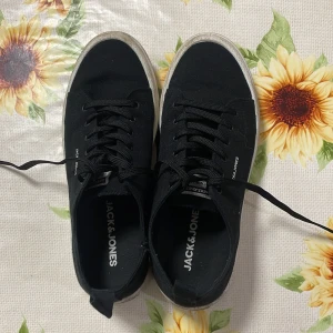 Svarta sneakers från Jack & Jones - Säljer ett par svarta sneakers från Jack & Jones i bra skick. De har en klassisk design med snörning och en vit sula som ger en snygg kontrast. Perfekta för vardagsbruk och passar till det mesta i garderoben.