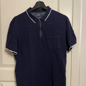 Mörkblå detaljerad pike stl L dressmann - Mörkblå kortärmad zip pike med vita detaljer från dressmann. Pilen bär stl L och är använd ett fåtal gånger men är helt ny i skick. Den har legat i en låda så är lite skrynklig men det går såklart att stryka.