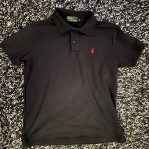 Svart pikétröja från Ralph Lauren - Säljer en klassisk svart pikétröja från Ralph Lauren i storlek M. Tröjan har en röd broderad logga på bröstet och är i mycket bra skick. Perfekt för både vardag och fest!