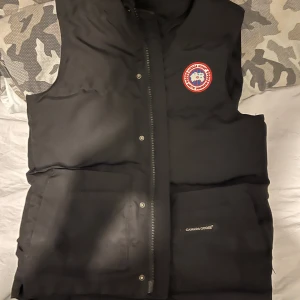 Canada goose väsy - Säljer en svart dunväst från Canada Goose i mycket bra skick. Västen har en dragkedja och knappar framtill samt ett snyggt märke på bröstet. Perfekt för höst och vinter, håller dig varm och stilren. Passar både till vardags och lite finare tillfällen. Storlek S
