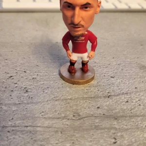 Fotbollsfigur Zlatan Ibrahimovic Manchester United  - En cool liten fotbollsfigur med röd tröja och vita shorts. Figuren står på en rund bas och har detaljer som svarta strumpor och röda skor. Perfekt för fotbollsfans som gillar att samla på miniatyrer!