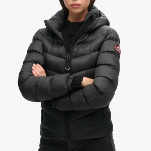 Svart dunjacka från Superdry - Säljer en svart dunjacka från Superdry i mycket bra skick. Jackan har en dragkedja framtill och en mysig huva. Perfekt för kalla vinterdagar med sin pufferstil och långa ärmar. Den är både snygg och praktisk för vintern!