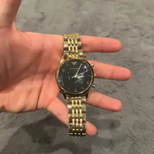 Säljer en stilren guldfärgad klocka från Emporio Armani. Den har en svart urtavla med guldiga siffror och visare. Armbandet är i rostfritt stål med en elegant länkdesign. Klockan kommer i originalförpackning med instruktioner. Perfekt för både vardag och fest! Köpt för 5000kr. Referens: AR1893