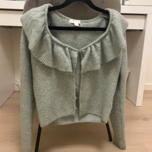 Ljusgrön stickad kofta med volangdetalj - Säljer en superfin stickad kofta med volangdetalj runt halsen. Den har långa ärmar och knappar framtill. Perfekt för höst och vinter när du vill vara både varm och stilren. Passar lika bra till vardags som till festligare tillfällen. 🧶✨