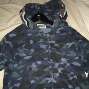 Camouflage hoodie från A Bathing Ape - Säljer en snygg hoodie från A Bathing Ape i blått camouflage-mönster. Den har en cool hajdesign på huvan och bokstäverna WGM i rött, blått och orange. Perfekt för streetwear-stilen med dragkedja framtill. Passar perfekt för höst och vår!
