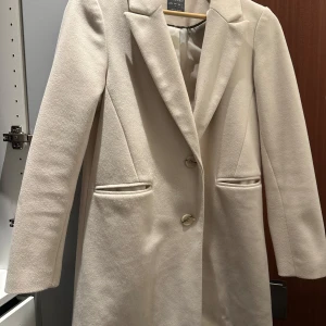 Beige kappa - Säljer en elegant beige kappa i mycket bra skick. Den har en klassisk design med knappar framtill och långa ärmar. Perfekt för höst och vår, och ger en stilren look till vilken outfit som helst.