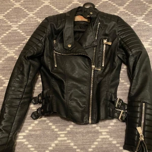 skinnjacka  - Säljer en snygg svart skinnjacka i nytt skick, aldrig använd. Den har dragkedjor och bältesdetaljer som ger en edgy look. Perfekt för höst och vår.