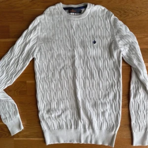 Vit stickad tröja från Morris - Säljer en stilren vit stickad tröja från Morris. Den har ett klassiskt flätmönster och en liten broderad logga på bröstet. Perfekt för både höst och vinter, med långa ärmar och ribbade kanter för extra komfort. Passar lika bra till vardags som till lite finare tillfällen.