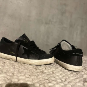 Svarta sneakers från Golden Goose - Säljer ett par svarta Golden Goose sneakers i bra skick. De har en cool stjärndetalj på sidan och klassisk snörning. Perfekta för både vardag och fest! Sulan är vit med en lätt sliten look som ger dem en unik stil.
