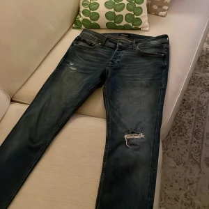 Mörkblå jeans med slitningar - Säljer ett par mörkblå jeans från Jack & Jones i storlek 32/32. De har en cool slitning på ena benet och en klassisk femficksdesign. Perfekta för en avslappnad stil och passar till både vardag och fest. Skicket är bra, men med en medveten sliten look. Kontakta via DM vid frågor!