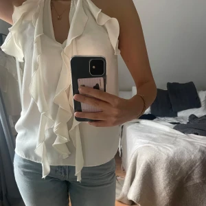 Zaratopp med  volangdetaljer - Säljer en elegant vit blus med volangdetaljer framtill. Den är ärmlös och har en stilren design som passar perfekt för både fest och vardag. Blusen är i mycket bra skick och med prislappen kvar!