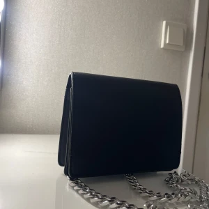 Svart axelväska med kedja - Snygg svart axelväska i skinn med en stilren design. Väskan har en silverfärgad kedja som axelrem, vilket ger den en modern touch. Perfekt för både vardag och fest!
