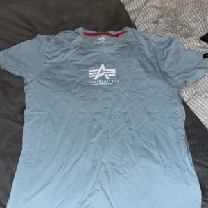 Turkos t-shirt från Alpha Industries - Säljer en snygg blå t-shirt från Alpha Industries i storlek M. Den har en vit logga tryckt på framsidan och en röd kant vid halsen. Perfekt för vardagsbruk eller avslappnade tillfällen. T-shirten är i utmärkt skick utan några tecken på användning. Den är gjord av mjukt bomullsmaterial.