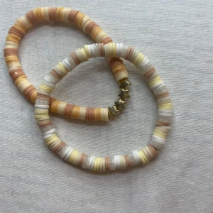 Färgglada armband och halsband - Två färgglada armband med platta pärlor i nyanser av beige, vit och gul. Ett av armbanden har små stjärndetaljer. Perfekt för att liva upp vilken outfit som helst!
