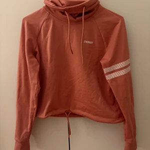 Rostfärgad hoodie från Stronger - Säljer en snygg rostfärgad hoodie från Stronger. Den har en hög krage och en randig detalj på ena ärmen. Den är i mycket bra skick och superbekväm att ha på sig. Passar perfekt till både träning och chill.