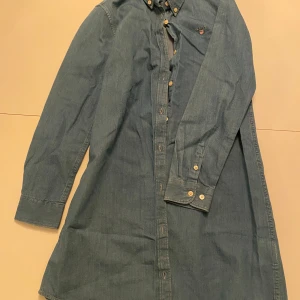 Blå jeansskjorta från GANT - Säljer en snygg blå jeansskjorta från GANT i storlek 146/152 cm, perfekt för 11-12 åringar. Skjortan har långa ärmar och knappar hela vägen ner. Den är i mycket bra skick och har en broderad GANT-logga på bröstet.