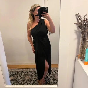 Svart oneshoulder klänning - Snygg svart oneshoulder klänning med en elegant slits på sidan. Perfekt för festliga tillfällen. Klänningen har en tight passform som framhäver figuren.Helt ny