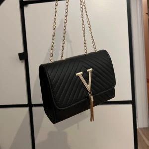 Svart quiltad axelväska med guldkedja - Snygg svart axelväska i quiltat mönster med en elegant guldkedja som axelrem. Väskan har en stilren design med ett stort V i guld på framsidan och en praktisk dragkedja inuti. Perfekt för både vardag och fest! Knappt använd, kanske 2 ggr max.