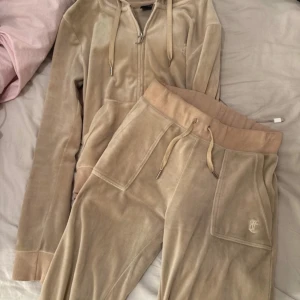 Beige velour hoodie och byxor från Juicy Couture - Säljer en supermjuk och bekväm beige velour hoodie och matchande byxor från Juicy Couture. Hoodien har dragkedja och snören, medan byxorna har en praktisk ficka med knapp. Perfekt för en avslappnad dag eller mysig hemmakväll. 🧡