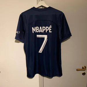 Blå PSG fotbollströja med Mbappé 7 - Säljer en snygg PSG fotbollströja i blått med Mbappé och nummer 7 på ryggen. Tröjan har korta ärmar och är i mycket bra skick. Den har vita och röda detaljer samt Qatar Airways som sponsorlogga på framsidan. Perfekt för alla PSG-fans!