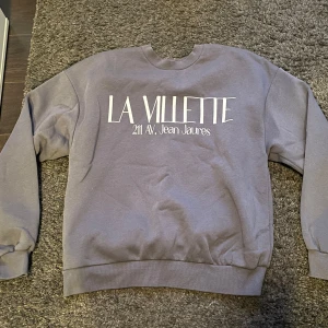 Grå sweatshirt med tryck - Säljer en mörkgrå sweatshirt med trycket 'LA VILLETTE 211 Av. Jean Jaures' på framsidan. Tröjan har en avslappnad passform och är perfekt för en casual look. Den är i bra skick och passar både till vardags och mysiga hemmakvällar. Den är ifrån Gina tricot. Använd nån enstaka gång men inget som syns. Superfin och bekväm tröja i storlek S.