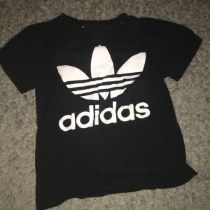 Svart t-shirt från Adidas - Säljer en svart t-shirt från Adidas i bra skick. Den har en stor vit Adidas-logga på framsidan. Perfekt för vardagsbruk eller sportaktiviteter. Passar barn i storlek 7-8 år.