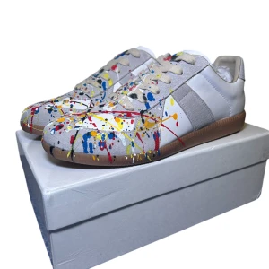 Maison Margiela Gat 43  - Säljer ett par unika sneakers från Maison Margiela i vitt skinn med ett coolt färgstänkmönster i rött, blått, gult och svart. Skorna har en klassisk sneakerdesign med snörning och en bekväm brun sula. Perfekta för att ge din outfit en konstnärlig touch! Allt på bilderna ingår. Skorna är helt nya