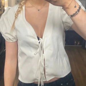 Vit blus med knytning - Säljer en söt vit blus med korta puffärmar och knytning framtill. Perfekt för sommardagar eller en avslappnad vårlook. Blusen har en v-ringad design och är i ett lätt och bekvämt material.