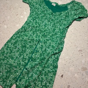 Grön blommig klänning från Pimkie - Säljer en superfin grön klänning från Pimkie med blommigt mönster. Den har korta ärmar och knappar framtill och knytning i bak. Perfekt för vår och sommar! 💚