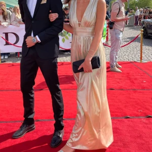 Champagnefärgad långklänning - Elegant champagnefärgad långklänning med djup v-ringning och ett brett midjeband som ger en smickrande siluett. Klänningen är perfekt för festliga tillfällen. Gamal balklänning, endast använd en gång! 