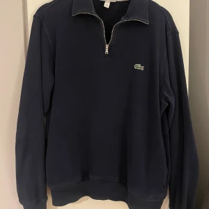 Mörkblå tröja från Lacoste - Säljer en snygg mörkblå half zip tröja från Lacoste. Tydligt använd och därför vissa slitningar på vissa ställen. Dock inget som syns