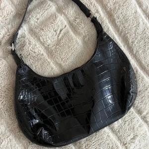 Svart axelväska i krokodilmönster - Snygg svart axelväska med krokodilmönster och dragkedja. Perfekt för en chic look, oavsett om du ska på fest eller bara vill ha en stilren accessoar till vardags. Väskan har en glansig finish som ger den en lyxig känsla.