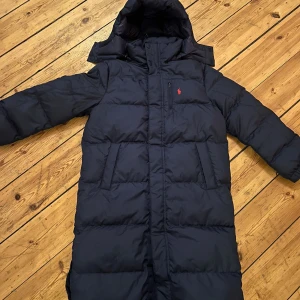 Mörkblå dunjacka från Polo Ralph Lauren - Säljer en snygg och varm mörkblå dunjacka från Polo Ralph Lauren. Jackan har en lång modell med huva och praktiska fickor. Perfekt för kalla vinterdagar. Den är i mycket bra skick och har en klassisk design med det ikoniska logotypen på bröstet.för barn 10-12 år 