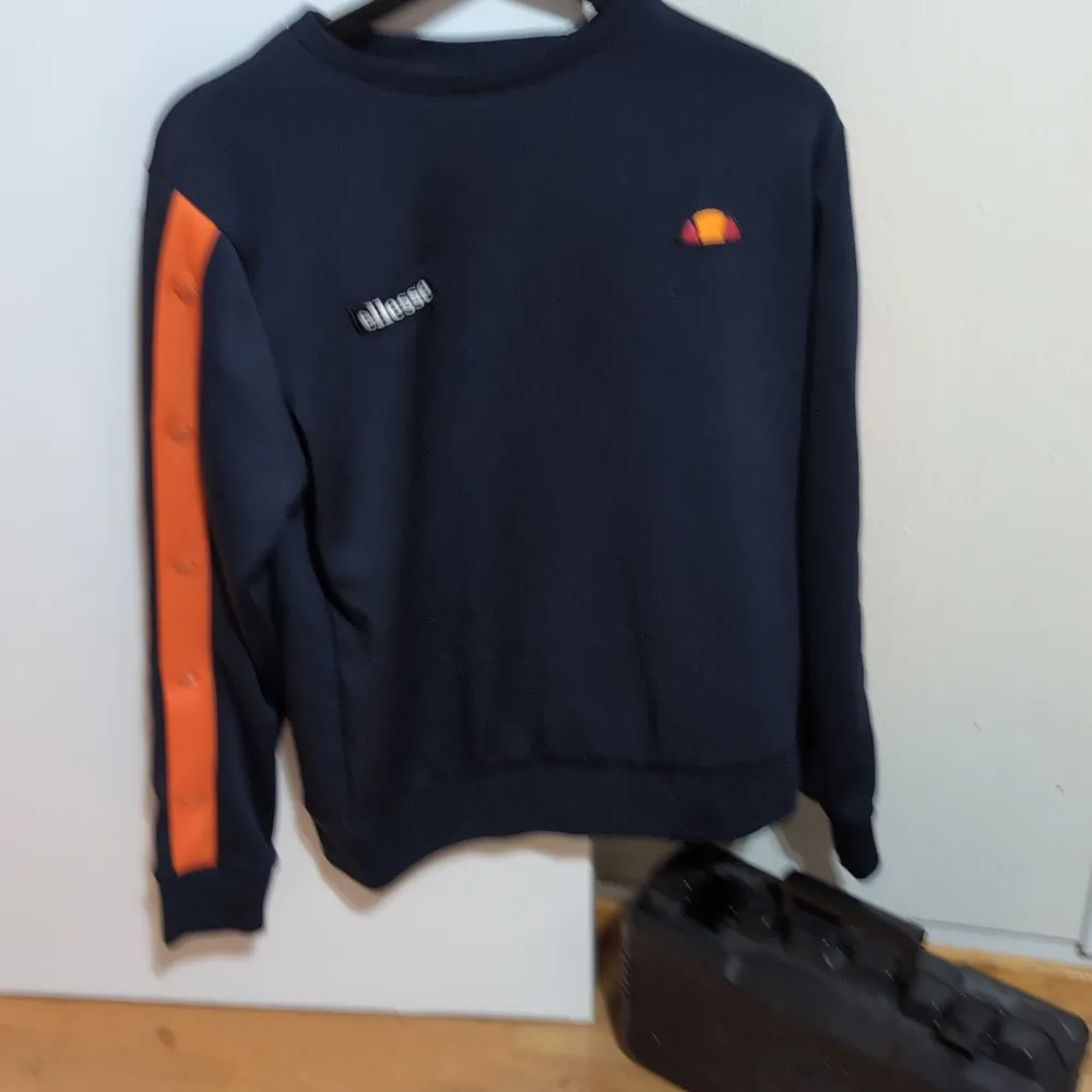 Säljer en snygg mörkblå tröja från Ellesse med orange detaljer på ärmarna. Perfekt för en casual look eller träning. Tröjan har en broderad logga på bröstet och är i bra skick. Passar perfekt till höst och vår!. Tröjor & Koftor.