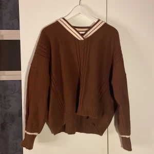 Brun stickad tröja med randig krage - Säljer en supermysig brun stickad tröja med en snygg vit och brun randig krage. Tröjan har långa ärmar och en avslappnad passform, perfekt för höst och vinter. Den har också ribbade muddar vid ärmslut och nederkant. Perfekt för att hålla sig varm och stilren!