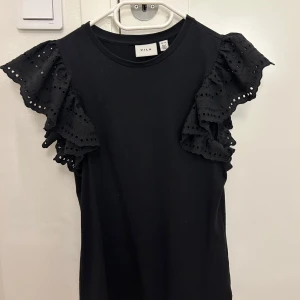 Svart topp med volangärmar från Vila - Säljer en snygg svart topp från Vila i storlek XS. Toppen har unika volangärmar med spetsdetaljer som ger en extra touch. Perfekt för både vardag och fest!