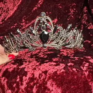 Både romantisk och dödligt vacker svart tiara. Känns verkligen retro i sin detaljerade utformning. Översållad med gnistrande svarta stenar och glittrande svart strass på en mycket mörkt guldfärgad botten.  Höjd fram 5,5, båglängd 32, bredd 14 cm. Ny.