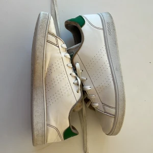 Vita Adidas sneakers - Säljer ett par vita Adidas sneakers med gröna detaljer. De har en klassisk design med snörning och perforerade sidor för ventilation. Skorna är lite smutsiga men i bra skick. Perfekta för vardagsbruk!